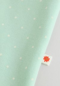 Helehall sinine kangas väikeste valgete täppidega ning väike valge silt, millel on punakasoranž lill kollase keskmikuga.