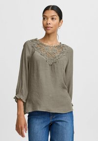 Blusa verde oliva con un colletto in crochet dettagliato, maniche lunghe con polsini a volant e una vestibilità comoda, realizzata in tessuto leggero.