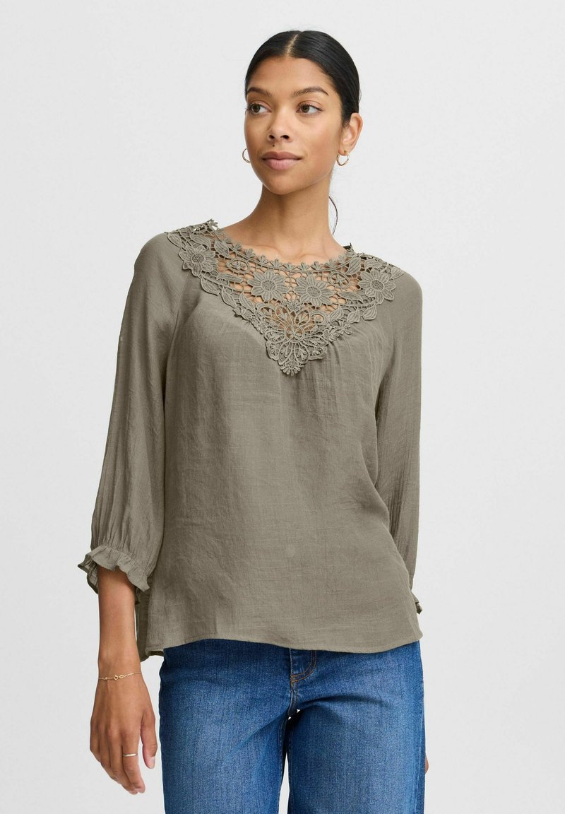 Blusa verde oliva con un colletto in crochet dettagliato, maniche lunghe con polsini a volant e una vestibilità comoda, realizzata in tessuto leggero.