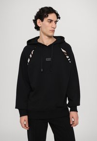Sweatshirt à capuche noir avec une coupe décontractée. Présente des accents latéraux rayés et une patch-logo rectangulaire sur le devant. Texture douce.