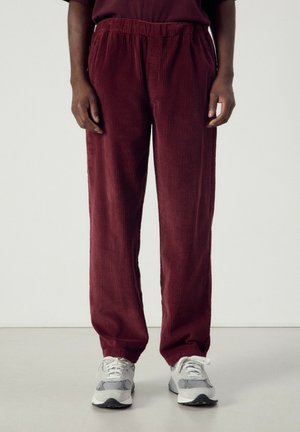 Persona che indossa pantaloni larghi di velluto a coste bordò e sneaker bianche, in piedi davanti a uno sfondo chiaro e uniforme.