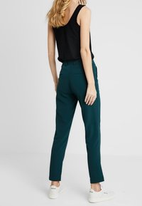 Svart linne och teal-byxor, åtsittande design, slät textur, rak benform, med vita sneakers, sedd bakifrån mot en ljus bakgrund.