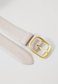 Vaalean beige nahkavyö, jossa on teksturoitu pinta, neliömäinen kultainen solki ja kohokuvioitu logo, litteä muotoilu ja terävä kärki.
