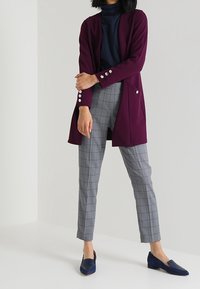 Osoba ubrana w ciemnofioletowy długi żakiet, granatowy top, szare kraciaste spodnie i granatowe mokasyny, stojąca na tle jednolitym.