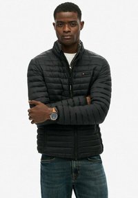 Czarna pikowana kurtka z zapięciem na zamek i stojącym kołnierzem. Posiada poziome przeszycia i naszywkę z logo. Noszona na białej koszuli i dżinsach.