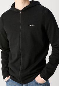 Schwarze Zip-Hoodie aus strukturiertem Stoff. Mit einem Logo auf der Brust, gerippten Akzenten an den Schultern und einer Kapuze mit Kordeln.