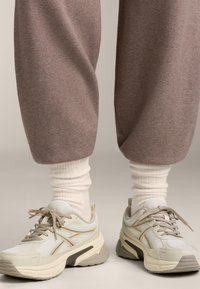 Hvide sneakers med beige accenter, mesh- og lædermaterialer, tykke såler og snørebånd. Varmtonede, løstsiddende grå træningsbukser og hvide sokker.