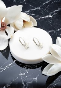 Boucles d'oreilles en argent en forme de fleur avec une pierre centrale, placées sur un présentoir rond blanc contre un fond en marbre noir et des fleurs blanches.