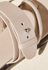 Lys beige sandaler med glatte syntetiske stropper og en metallisk nitte detalje. Har en flad sål og et minimalistisk design.