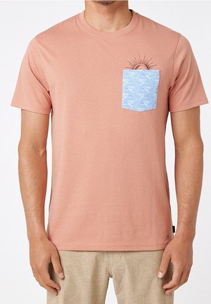 Mężczyzna w łososiowo-różowej koszulce z jasnoniebieską kieszenią ozdobioną białym wzorem fal i grafiką słońca częściowo za kieszenią.