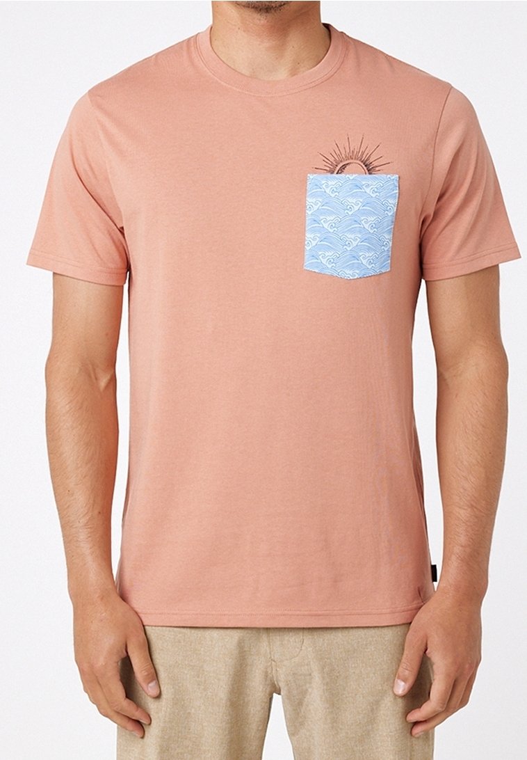 Homme portant un t-shirt saumon avec une poche bleu clair décorée de motifs de vagues blanches et un graphique de soleil partiellement derrière la poche.