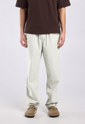 Homme portant un pantalon beige, une chemise marron à manches courtes et des chaussures en daim marron clair sans lacets, debout devant un fond uni.