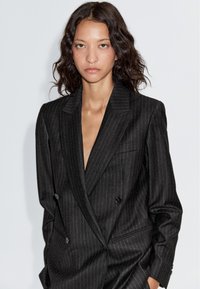 Zwarte blazer met krijtstreep, dubbelgeknoopt ontwerp, pieklapels en een subtiele structuur. Voorzien van zijzakken en knoopdetails.