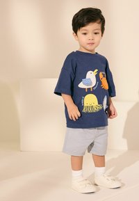 Marinblå bomullströja med lekfulla havsdjursgrafik; matchad med ljusgrå shorts och vita slip-on skor med två remmar och ribbade strumpor.