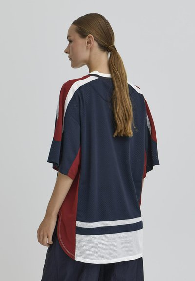 Femme aux longs cheveux bruns en queue de cheval, portant un maillot de sport ample à manches courtes avec des blocs de couleurs bleu marine, rouge et blanc, se tenant de profil.