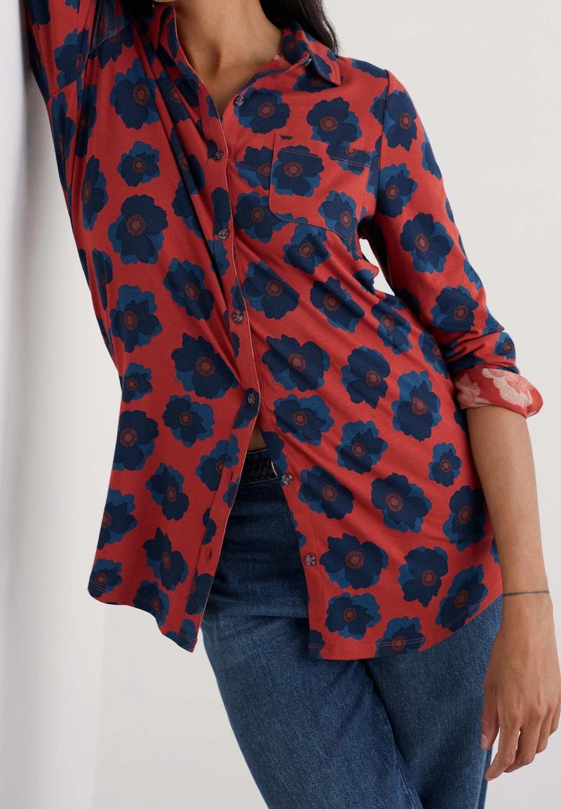 Femme portant une chemise rouge à boutons avec un grand imprimé floral bleu foncé et des manches retroussées, associée à un jean bleu, appuyée contre un mur.