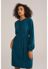 Turkos klänning i mjukt tyg, med långa ärmar, rund halsringning och midja med bälte och knytdetalj fram.