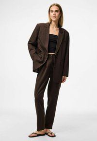 Bruin op maat gemaakt pak met een lange blazer met een enkele knop en zakdetails, gecombineerd met rechte broekspijpen, gedragen over een zwarte top.