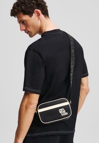 Sac bandoulière noir avec finition texturée, accents crème et logo. La bandoulière réglable arbore un motif monogramme. Forme rectangulaire simple.