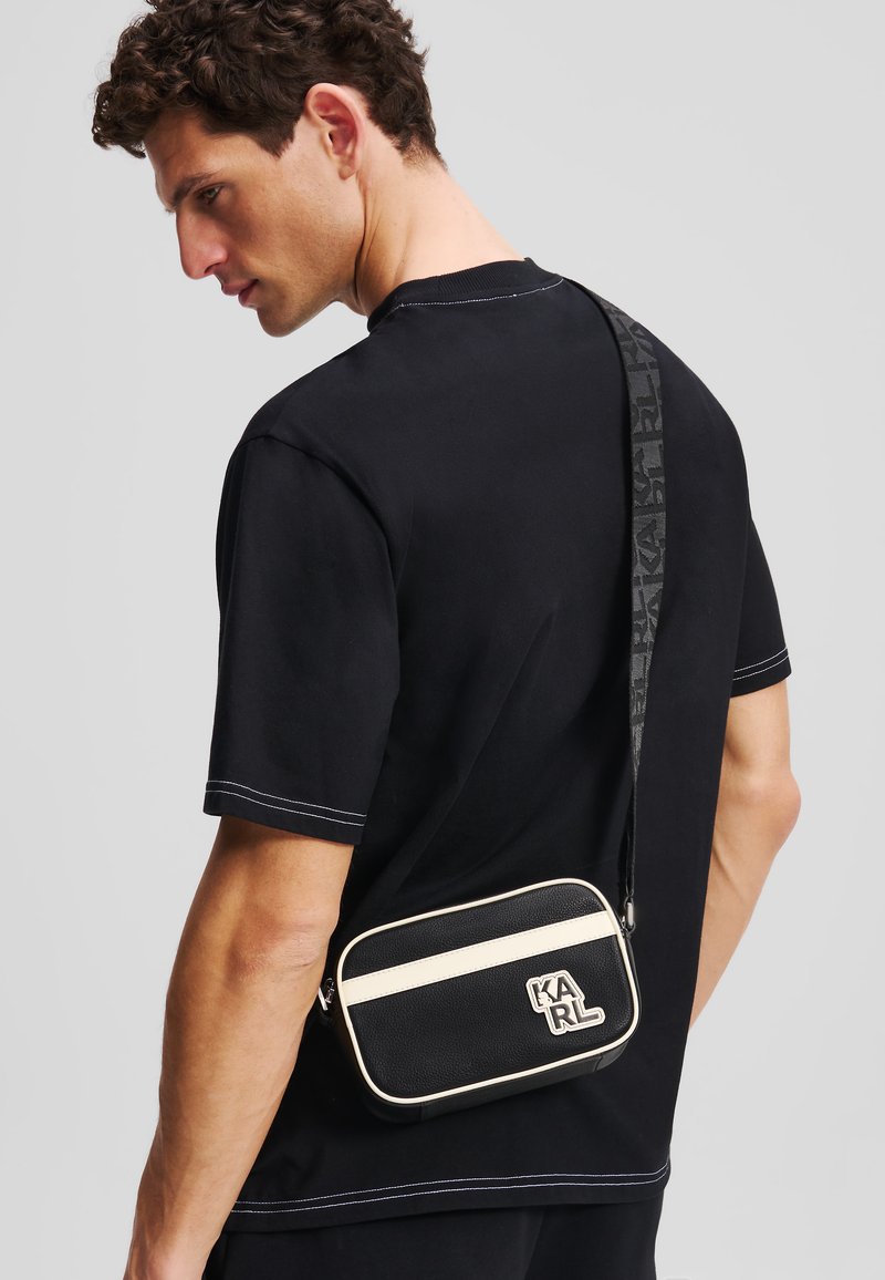 Sac bandoulière noir avec finition texturée, accents crème et logo. La bandoulière réglable arbore un motif monogramme. Forme rectangulaire simple.