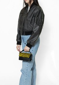 Giacca bomber in pelle nera con polsini a coste, indossata sopra un denim azzurro chiaro. Tiene una piccola borsa con una patta a stampa leopardata gialla e base nera.