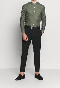 Miesten oliivinvihreä pitkähihainen paita, jossa on kaulus ja napitettava etu, yhdistetty mustiin kapealahkeisiin housuihin ja mustiin loafer-kenkiin.