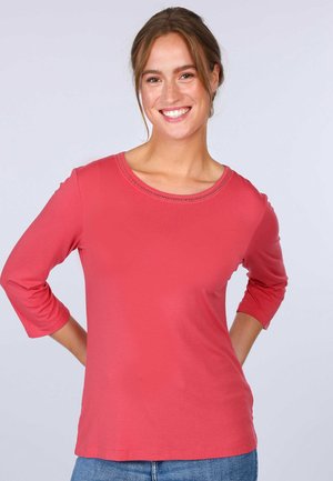 Coral long-sleeve top with a round neckline and decorative stitching. Soft fabric with a smooth texture. Paired with denim jeans.
