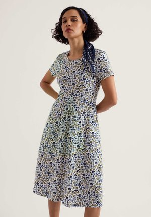 Žena s tamnom kovrčavom kosom, odjevena u cvjetnu haljinu dužine do koljena s kratkim rukavima i mornarskoplavi šal vezan oko glave.