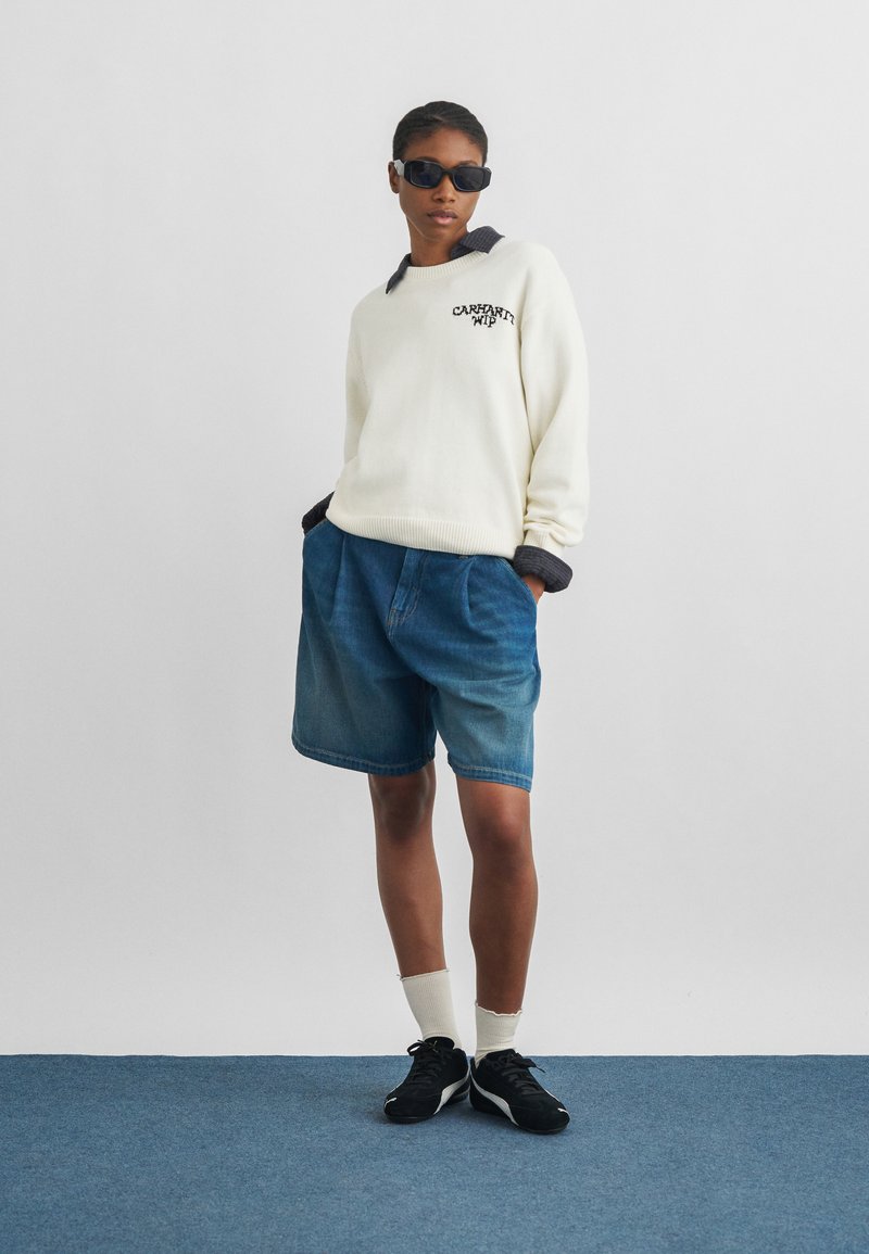 Persona con occhiali da sole, maglione bianco Carhartt WIP, pantaloncini di jeans blu, calzini bianchi e scarpe da ginnastica nere in piedi su un tappeto blu.