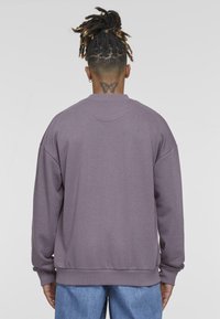 Felpa oversize viola con colletto e polsini a costine, caratterizzata da una texture morbida. Sul retro presenta un tatuaggio di farfalla appena sopra lo scollo.