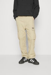 Pantaloni cargo beige con numerose tasche grandi, vestibilità rilassata e cuciture a vista. Abbinati a sneaker nere con strisce bianche.