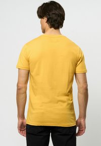 Tricou galben din bumbac cu mâneci scurte, decolteu rotund și o textură netedă. Îmbrăcămintea este asociată cu pantaloni negri, punând accent pe vederea din spate a tricoului.