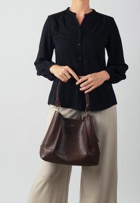 Bolso de mano de cuero marrón con un diseño estructurado, base redondeada y asas cortas. El bolso cuenta con un panel frontal decorativo y detalles en tono dorado.