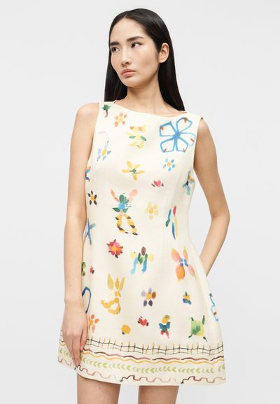 Γυναίκα που φοράει λευκό φόρεμα χωρίς μανίκια με πολύχρωμα αφηρημένα λουλουδάτα σχέδια, στέκεται μπροστά σε απλό φωτεινό φόντο.