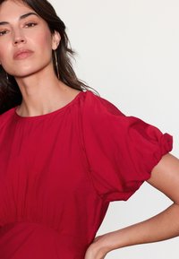 Tummanpunainen pusero, jossa on liehupusuhihat ja pyöreä pääntie. Vyötäröllä on kerrostettu kuviointi ja kangas on sileä.