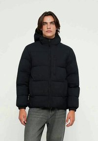 Casaco preto acolchoado com design almofadado, fecho de correr e capuz. Apresenta um bolso no peito e punhos elásticos. Usado com jeans cinzentos.