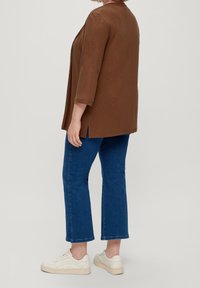 Cardigan marron à manches longues avec des fentes latérales, associé à un jean court bleu et des baskets blanches, vu de dos.