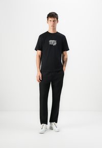 Black cotton t-shirt with a graphic print on the chest, paired with black trousers and white sneakers. Simple, classic design.