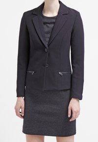 Granatowa marynarka z klapami, dwoma guzikami i zamykanymi kieszeniami. Noszona na tle strukturalnej, ciemnoszarej sukienki. Dopasowany krój i gładki materiał.