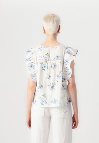 Top floreale bianco con fiori blu, maniche a balze e dettagli in pizzo lungo la schiena. Tessuto leggero e traspirante.