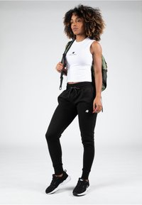 Zwarte sweatpants met een tailleband met trekkoord en zijstrepen. Witte mouwloze top met een logo. Zwarte sportschoenen en een groene rugzak.