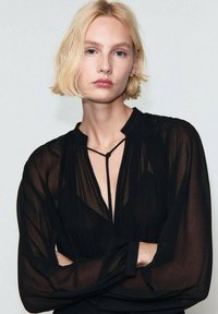 Zwarte transparante blouse met een V-hals, lange mouwen en een strikdetail bij de halslijn. Glad textuur en lichte plooien bij de schouders.