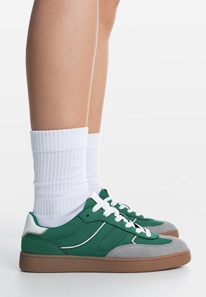 Piernas con calcetines blancos hasta el tobillo y zapatillas bajas verdes, grises y marrones con cordones blancos, de pie sobre una superficie blanca.
