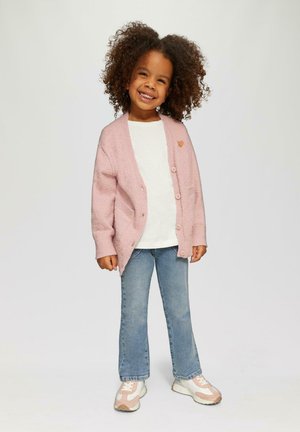 Glimlachend kind met krullend haar, gekleed in een roze cardigan, witte shirt, blauwe spijkerbroek en roze-witte sneakers, staand tegen een neutrale achtergrond.