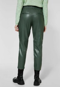 Vihreät tekonahkabehkeet, joissa on joustava vyötärö, kaksi takataskua, kapeneva lahje ja sileä, kiiltävä pinta. Mustat saappaat näkyvissä.