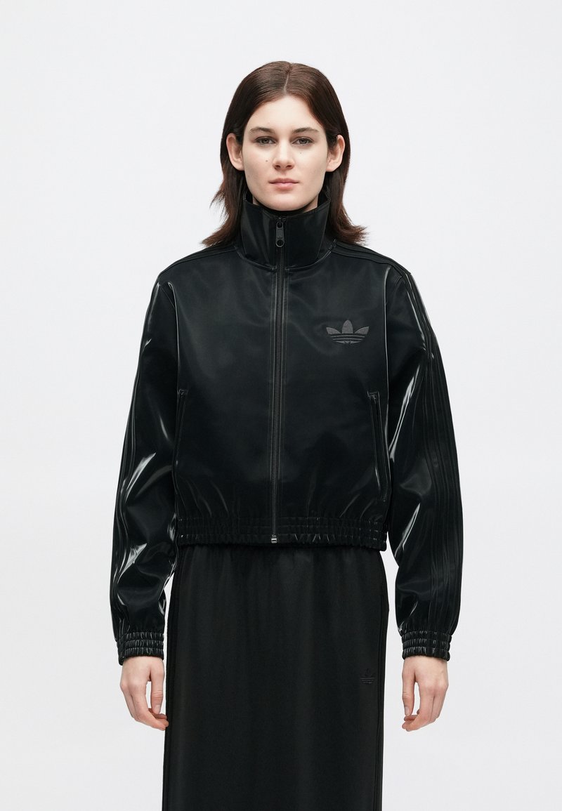 Person in einer schwarzen, mit Reißverschluss zu schließenden Jacke mit hohem Kragen und Logo auf der Brust, kombiniert mit einem passenden schwarzen Rock, steht vor einem schlichten Hintergrund.