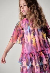 Farverig, lagdelt kjole i gennemsigtigt stof med flæsekant og korte ærmer; indeholder livlige mønstre i pink, lilla og gule nuancer.