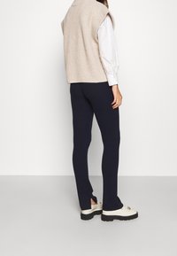 Beige gestrickter ärmelloser Pullover über einem weißen Hemd, kombiniert mit marineblauen gerippten Leggings und weißen Schuhen mit einer dicken schwarzen Sohle.