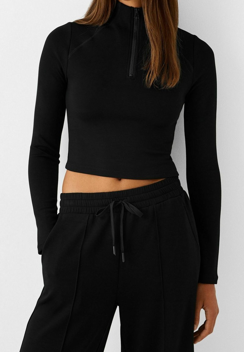 Haut noir court à manches longues avec col montant et fermeture éclair à moitié, associé à un pantalon de jogging noir à taille élastique avec poches latérales.