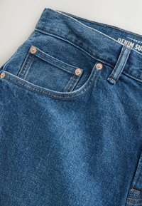 Tuvplāns zilās džinsa biksēs, redzamas šuves, kniedes, jostas cilpa un daļēja zīmola etiķete jostas vietā.